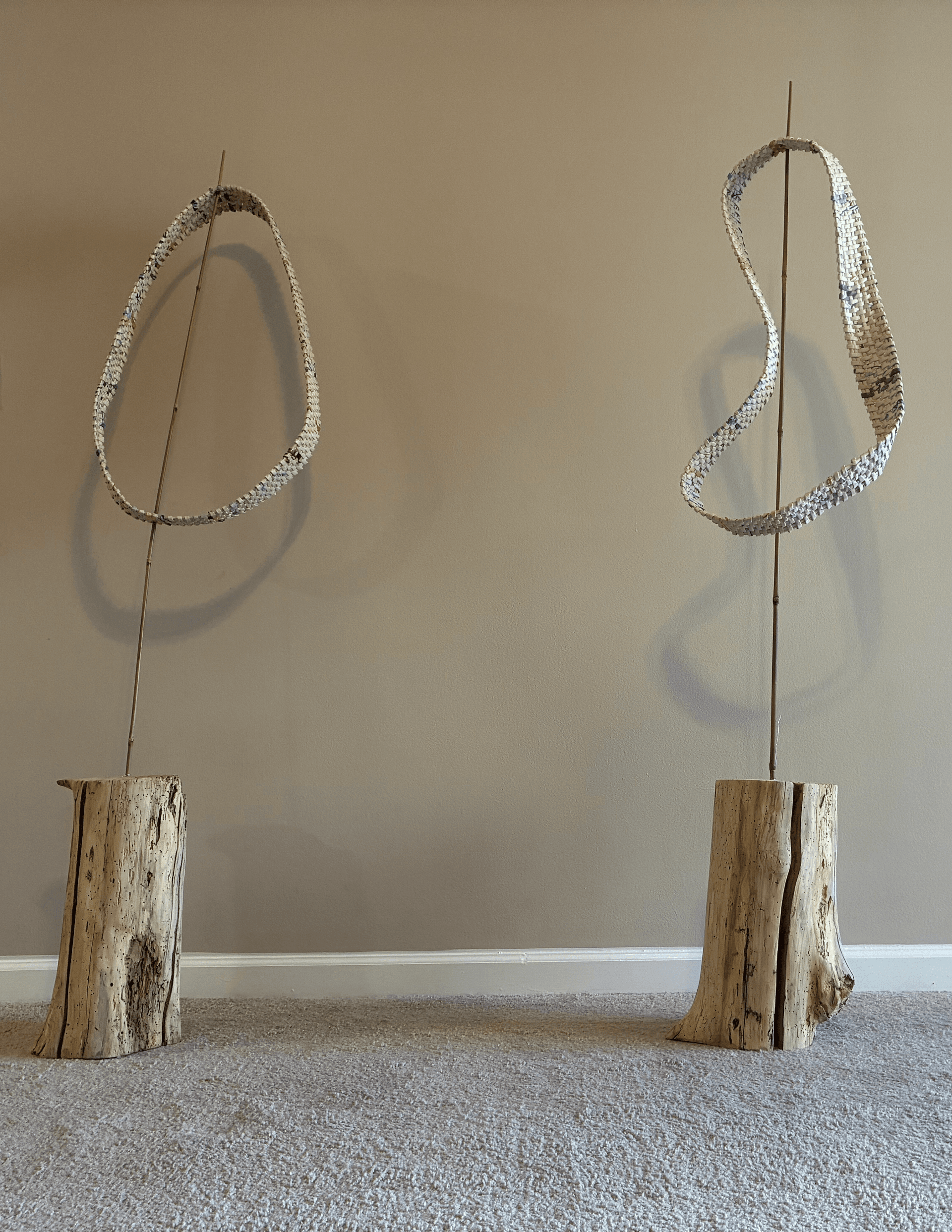 Woven loop sculpture made from rolled paper strips, twisting in a continuous band with multiple rotations, mounted on a weathered wood base with bamboo supports.