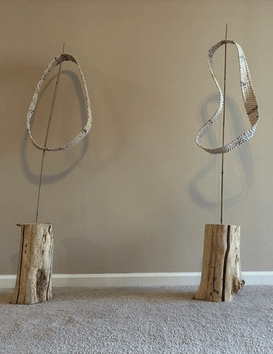 Woven loop sculpture made from rolled paper strips, twisting in a continuous band with multiple rotations, mounted on a weathered wood base with bamboo supports.
