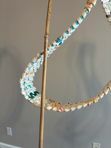 Woven loop sculpture made from rolled paper strips, twisting in a continuous band with multiple rotations, mounted on a weathered wood base with bamboo supports.