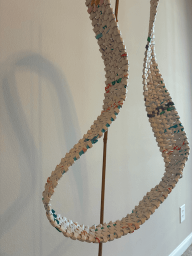 Woven loop sculpture made from rolled paper strips, twisting in a continuous band with multiple rotations, mounted on a weathered wood base with bamboo supports.