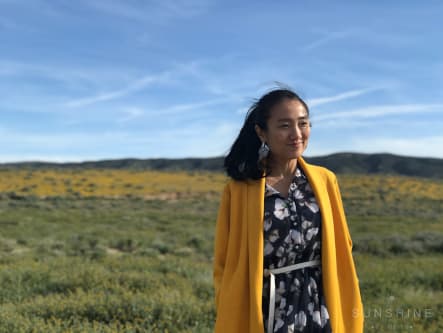 Clover Xuesong Zhou in a long yellow coat standing in a windy field of yellow wildflowers.
